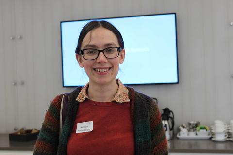 Photo of woman wearing red shirt, striped cardigan and dark-framed glasses in front of wall-mounted screen